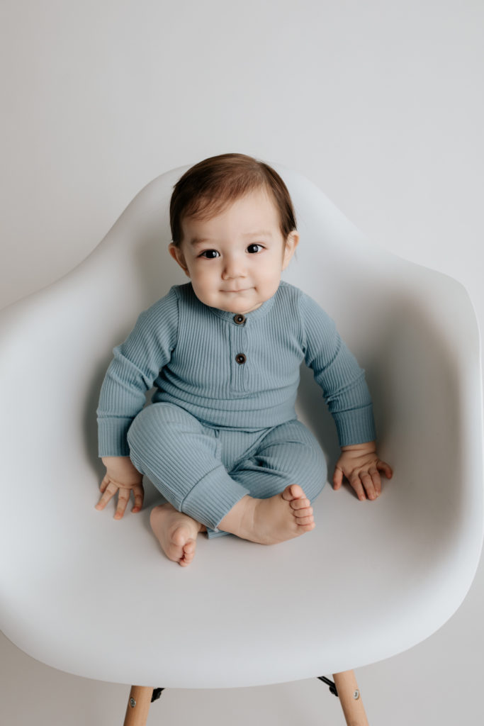 Baby boy wearing blue and sitting in a white chair during his cake smash portrait session.