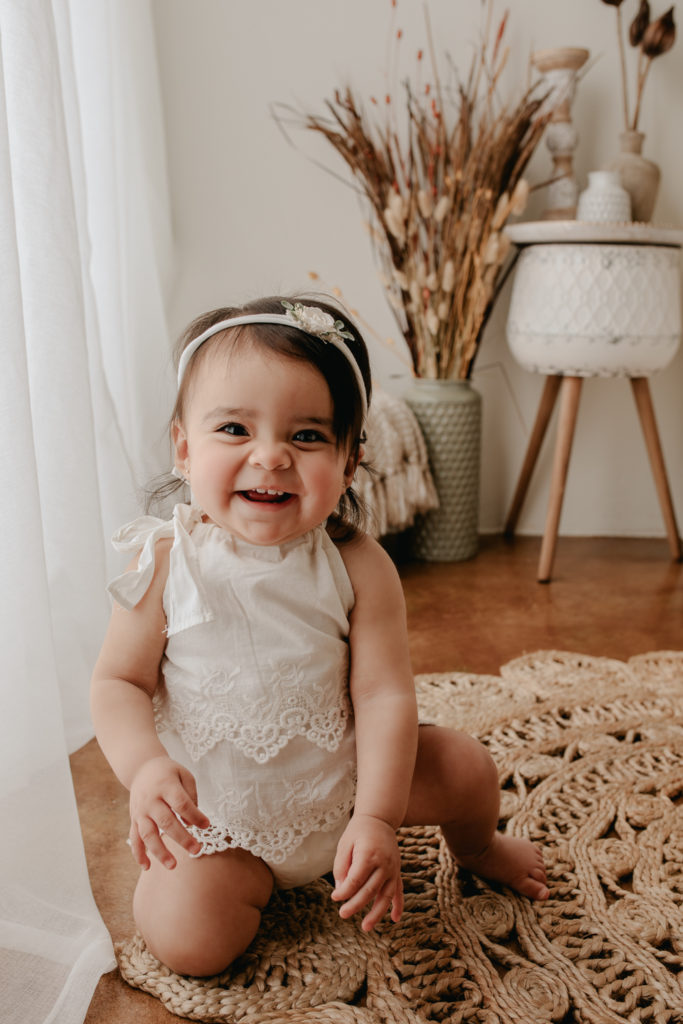Baby girl wearing white and sitting on the floor during his cake smash portrait session.
