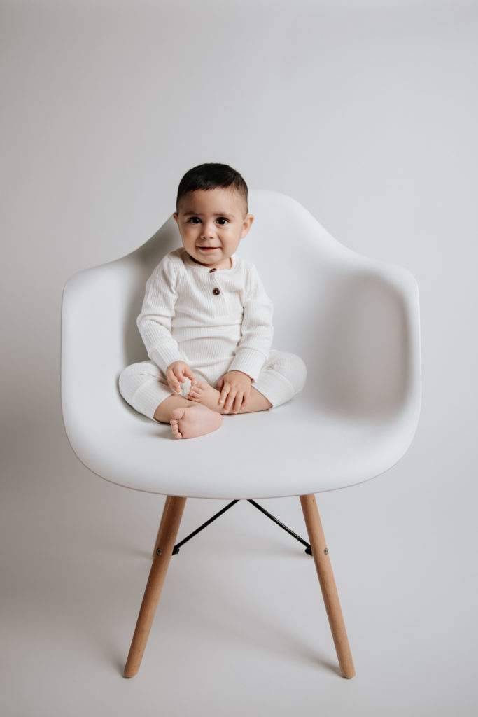 Baby boy wearing white and sitting in a white chair during his cake smash portrait session.
