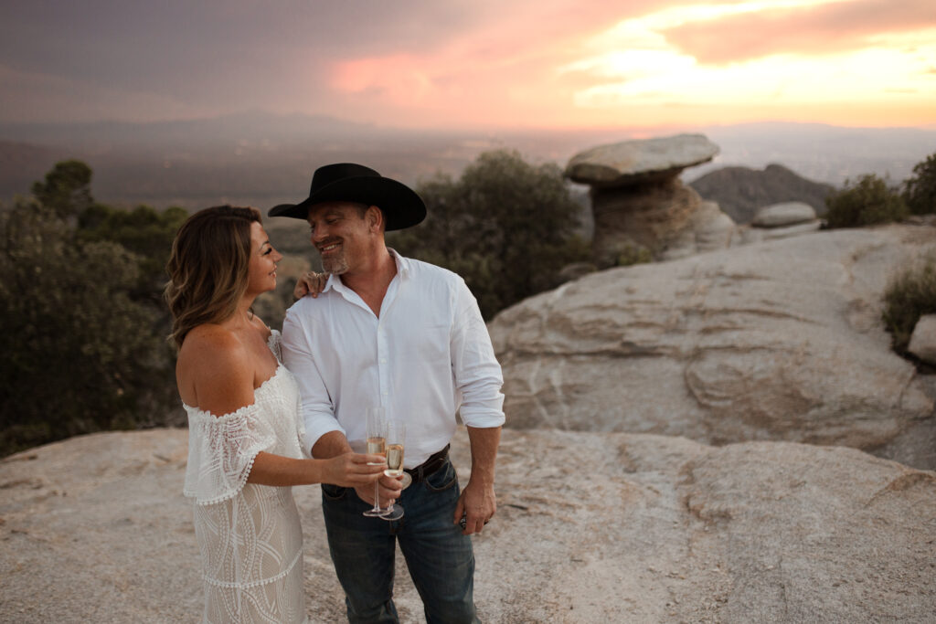 A cute couple sharing a glass of champagne to celebrate their Arizona elopement. They are on top of a mountain looking over Tucson with a pretty sunset in the backgound.