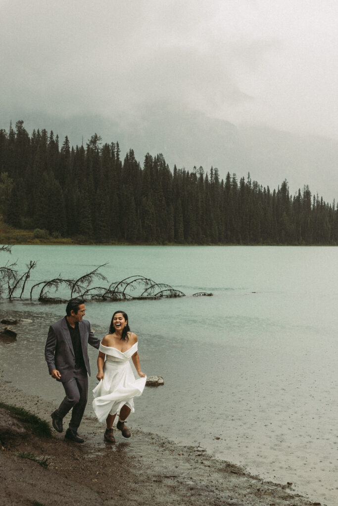 This newly wed couple is running along the lake shore. It is raining but they don't care. They are just happy to be eloping in Banff Nation Park.