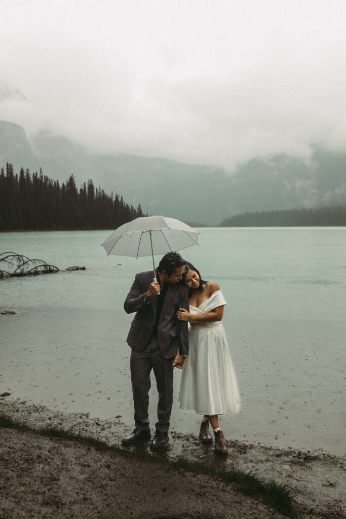 a man and his wife are standing on the lake shore under an umbrella. She is in her wedding dress and enjoying the moment after they eloped.