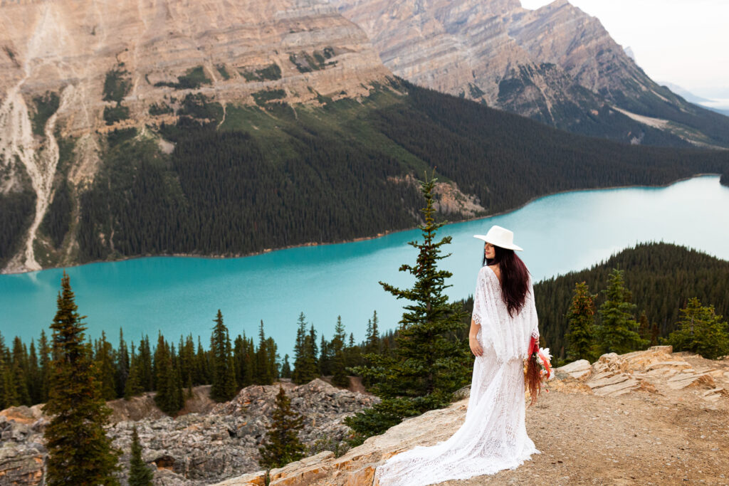 Beautiful bride stands above a lake ready for her Banff elopement. She is stunning in her white dress with the blue lake below.