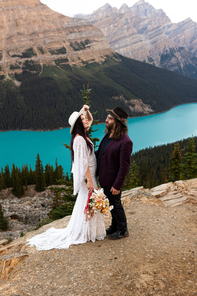 This is such a beautiful moment. the bride is enjoying the view from on top of a mountain looking down on the blue lake while her groom is admiring her. 