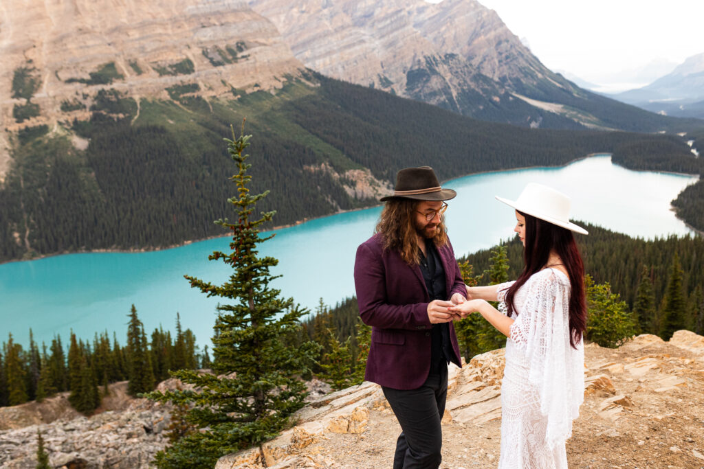 Groom is giving his bride her wedding ring. they are standing on top of a mountain ready to celebrate their Banff elopement.